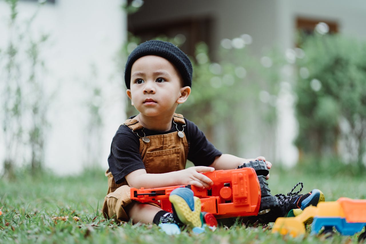 Cute boy in overalls playing with colorful toy trucks on the grass in a garden setting.