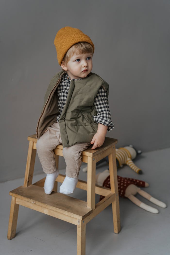 Cute child wearing beanie and winter clothes, sitting on wooden stool in indoor studio setting